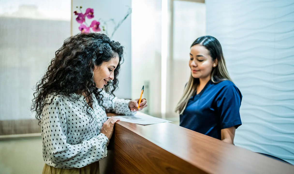 Patient filling out paperwork at a brightly lit medical clinic reception desk with a staff member in blue scrubs.